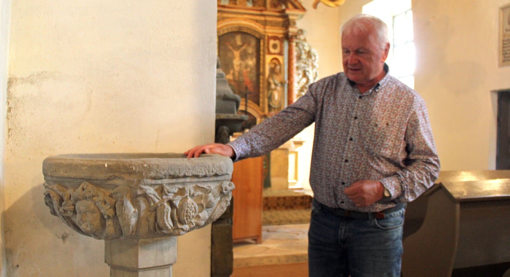 Gunther Hirschligau an dem 350 Jahre alten Taufstein in der Wormsdorfer Kirche. An der Wand befindet sich außerdem ein Gedenkstein, der an den Steinmetz Melchior Lentz erinnert.