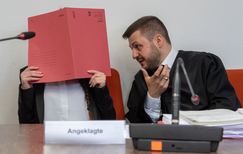 Die Angeklagte Jennifer W. hält im Gerichtssaal einen roten Aktendeckel vor das Gesicht. Rechts neben ihr sitzt Anwalt Ali Aydin.