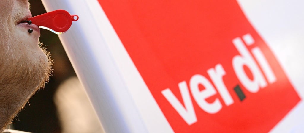 Ein Mann hat bei einem Warnstreik vor dem Verdi-Logo eine Pfeife im Mund.