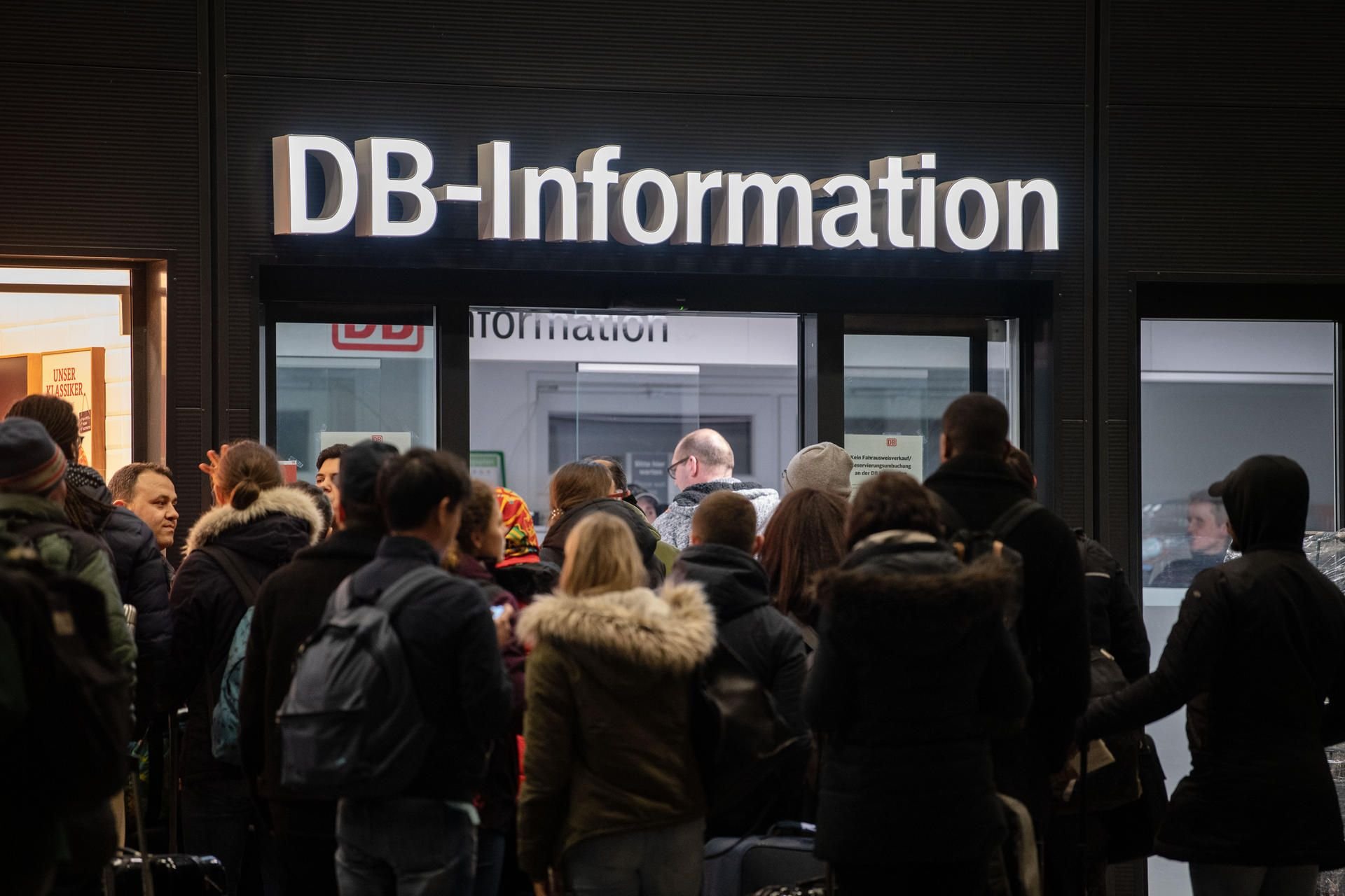 Reisende warten vor der Information der Deutschen Bahn in einem Bahnhofsgebäude.