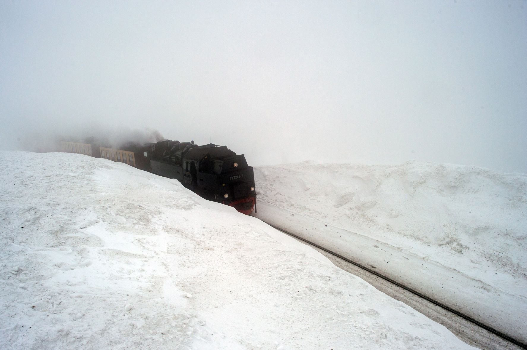 Ein Dampfzug der Brockenbahn fährt im Nebel zwischen zwei meterhohen Schneeverwehungen hindurch auf den Brockengipfel.