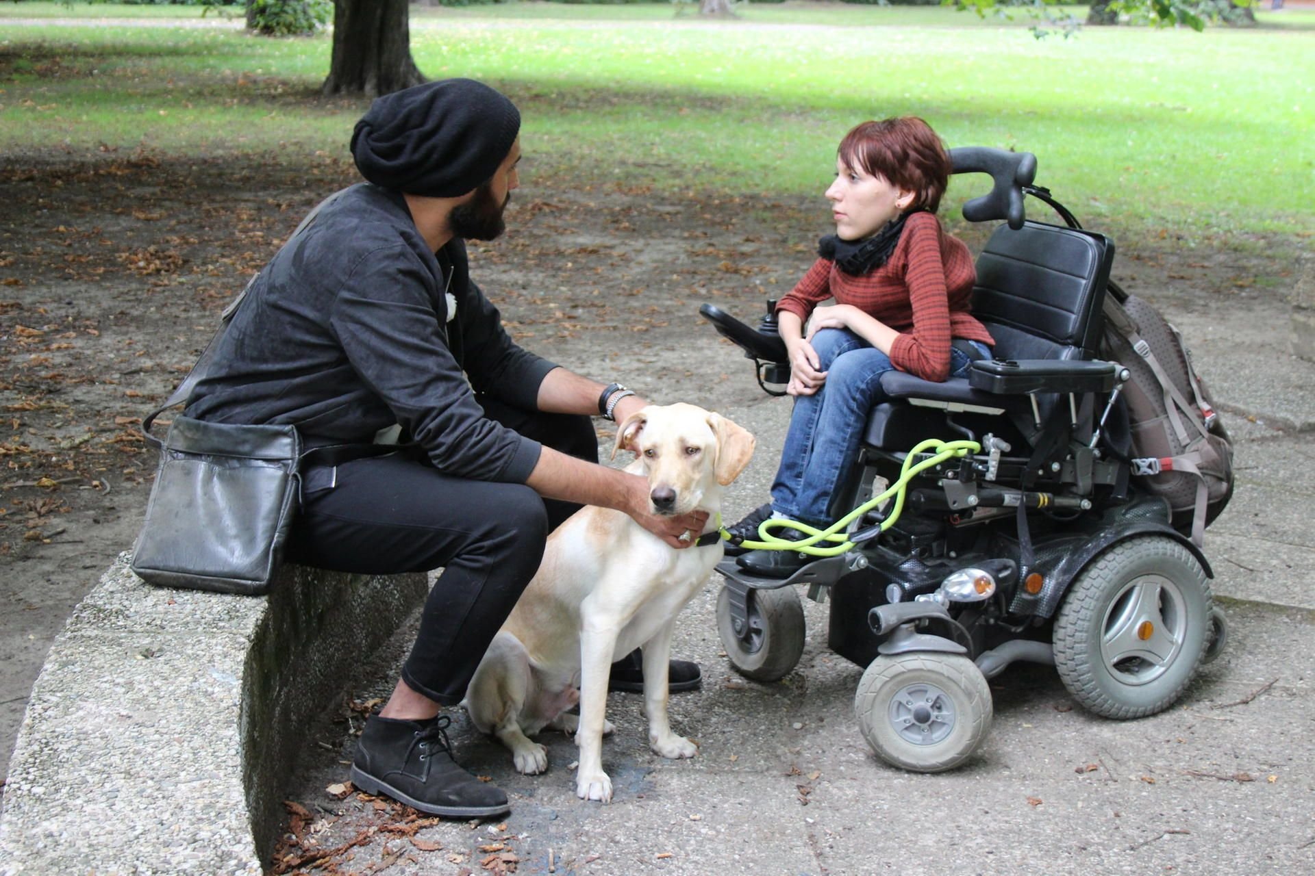Hundetrainer Masih Samin (l.) im Gespräsch mit Sarah aus Leipzig (r.), die im Rollstuhl sitzt und eine Teilnehmerin von „Projekt Superhund - Helfer auf vier Pfoten“ ist. Mittendrin: Hund Fito, den Samin zum Superhund trainieren möchte.