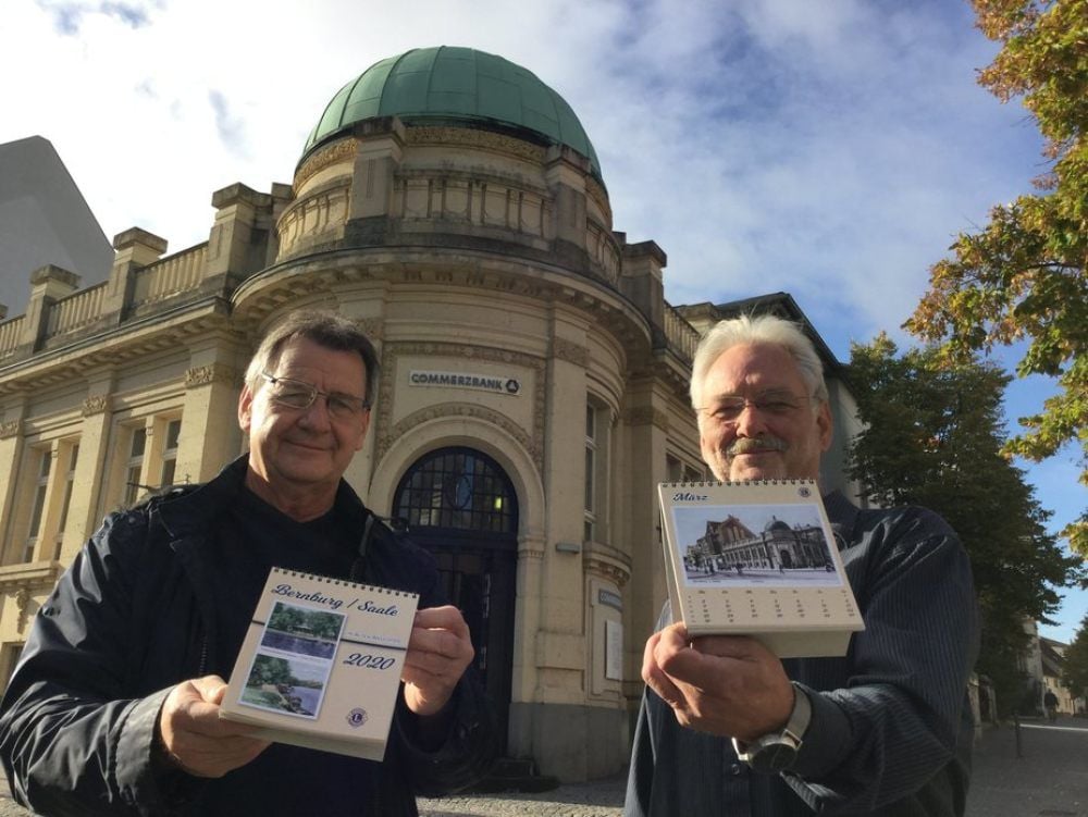 Winfried Becker (links) und Michael Koch präsentieren vor dem Eckgebäude Friedensallee/Poststraße, das sich in früherer Ansicht im März-Blatt wiederfindet, den Jahreskalender 2020 mit Motiven aus Bernburg.