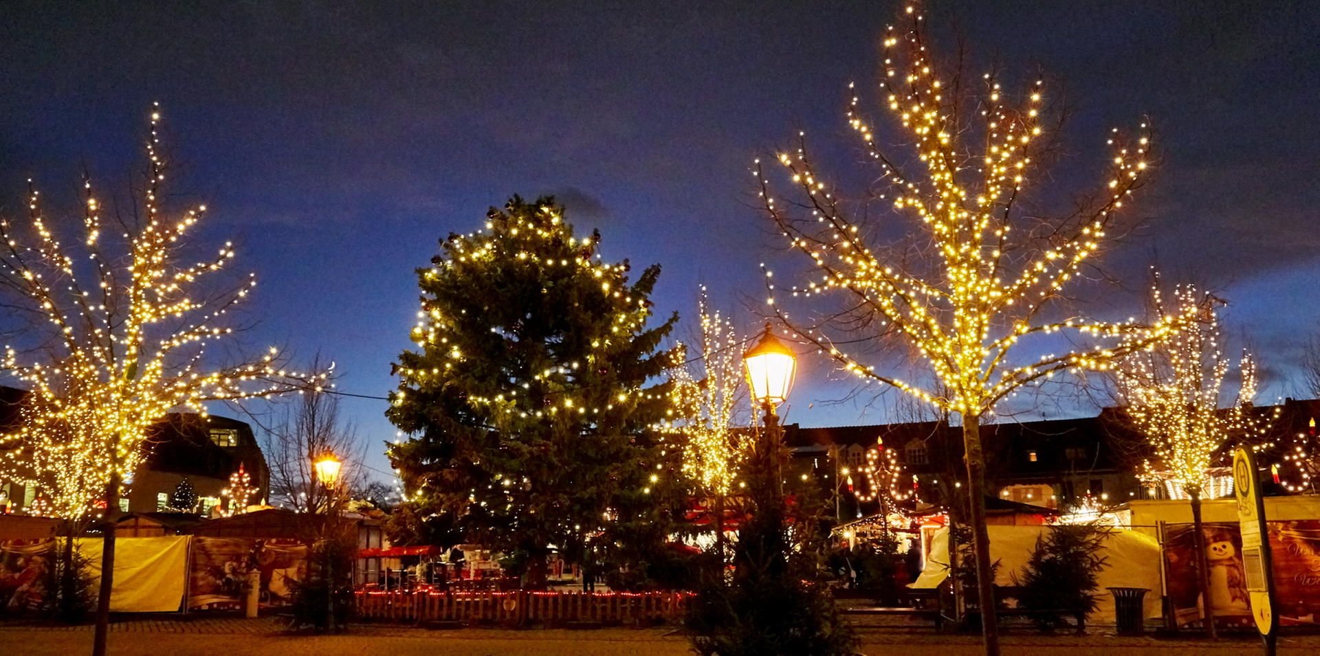 Lichterketten in den Linden am Rande des Weihnachtsmarktes sorgen für eine optische Wärme, auch Buden und Stehtische sind stimmungsvoll erleuchtet.