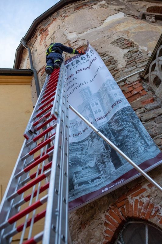Große Banner sollen am Schloss Droyßig für die Ausstellung „Lost Places“ werben. Die Feuerwehr, hier Patrick Winkler, hilft beim Anbringen.