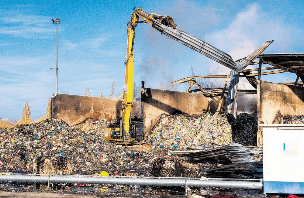 Der Spezialbagger nimmt auf dem Gelände im Industriegebiet Krumpa, wo seit Samstagnacht der Brand einer Lagerhalle mit Kunststoffmüll gelöscht wird, seine Arbeit auf.
