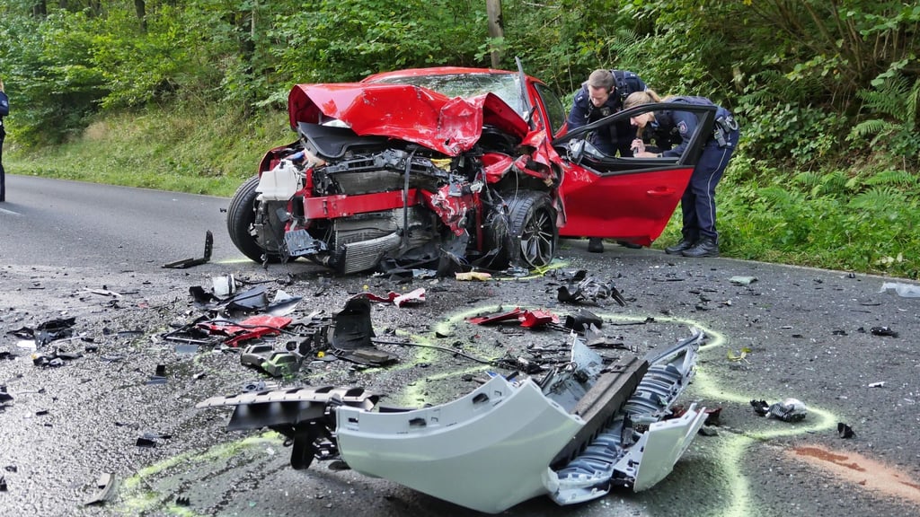 Polizisten stehen an einem zerstörten Auto auf einer Landstraße bei Siegen.