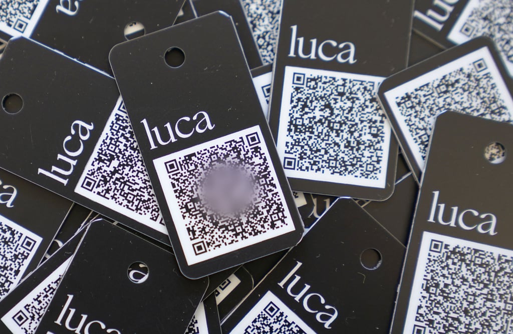 Illustration: Schlüsselanhänger mit QR-Code und der Aufschrift „luca“ liegen auf einem Tisch.