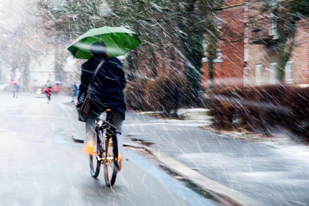 Ein Radfahrer mit Regenschirm in der Hand fährt durch Schneeregen.