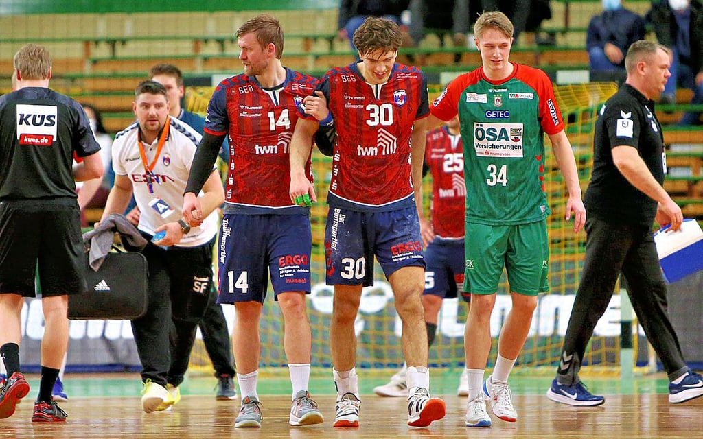 HCB-Spieler Marcel Popa (Nummer 30) hat sich am Knie verletzt und wird von seinem Teamkollegen Florian Pfeiffer und dem Magdeburger Justin Döbler, der für das vorherige Foul die Rote Karte sah, vom Spielfeld geführt.