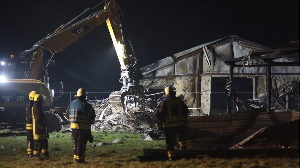 Einsatzkräfte der Feuerwehr stehen nach den Löscharbeiten neben einem Bagger, der Metallteile vor der niedergebrannten Hühnerstallanlage beseitigt.