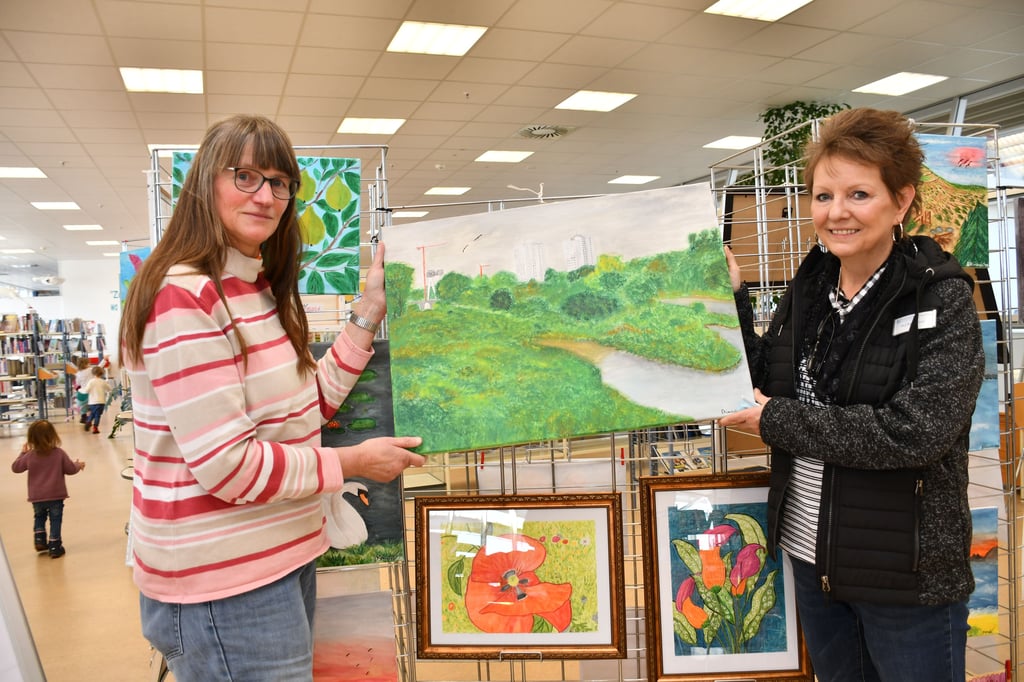 Gabriele Kaminski und Ilka Baake-Ernst zeigen eins der Bilder der Ausstellung in der Stadtteilbibliothek im Magdeburger Flora-Park.