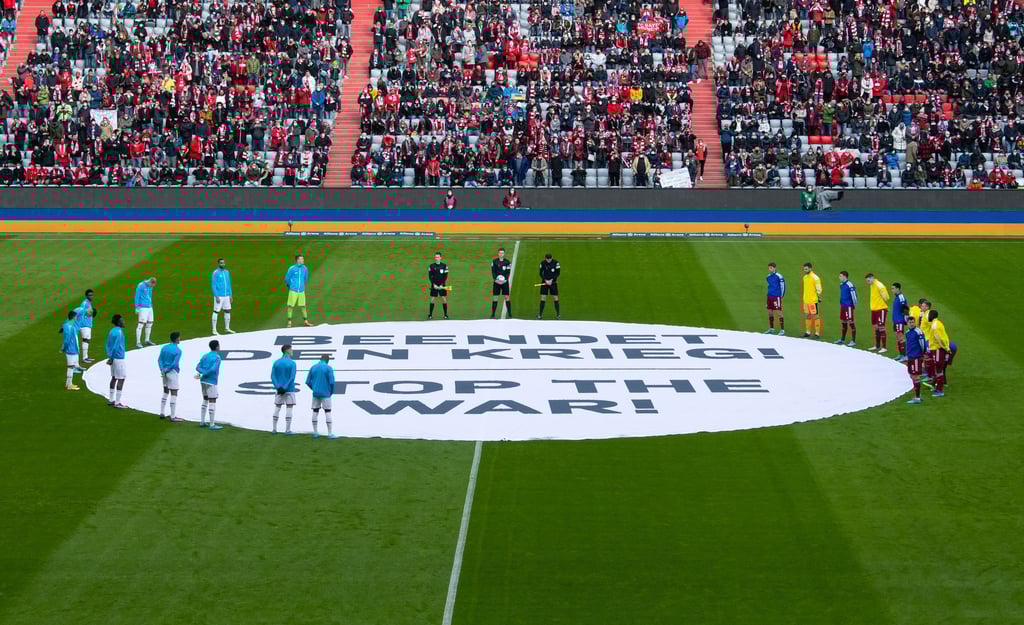 Die Spieler von München und von Leverkusen stehen um ein Banner mit „Beendet den Krieg! - Stop the War!“.