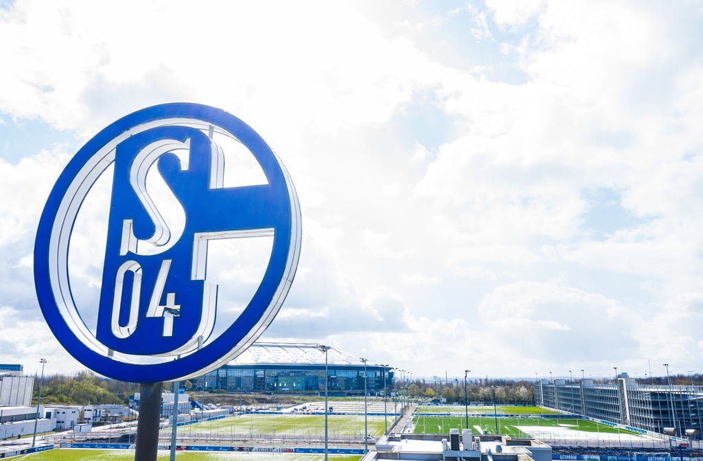 Im Vordergrund dreht sich das Logo des FC Schalke auf der Geschäftsstelle, im Hintergrund steht die Veltins Arena.