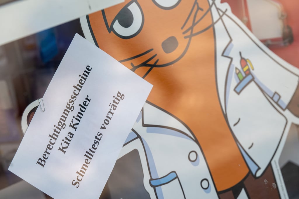 Ein Schild mit der Aufschrift „Berechtigungsscheine Kita Kinder - Schnelltests vorrätig“ hängt im Schaufenster einer Apotheke.