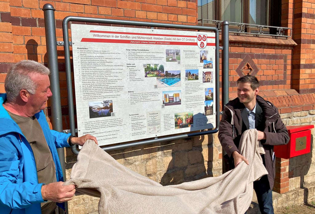 Ulrich Hartleb (links), Vorsitzender des Heimatvereins Alsleben, enthüllt mit Bürgermeister Alexander Siersleben die neue Infotafel vor dem Rathaus.