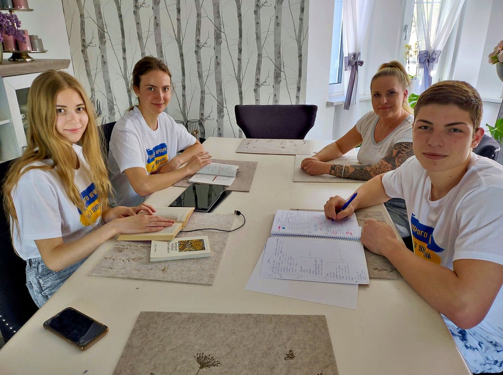 Hausaufgabenzeit mit Agnieszka Hinz (rechts hinten). Links am Tisch sitzen Sasha und ihre Schwester Ksenja (von links), vorne rechts befindet sich Kirill. Alle drei tragen T-Shirts mit den Farben der Ukraine und dem Spruch „Guten Abend, wir kommen aus der Ukraine“.