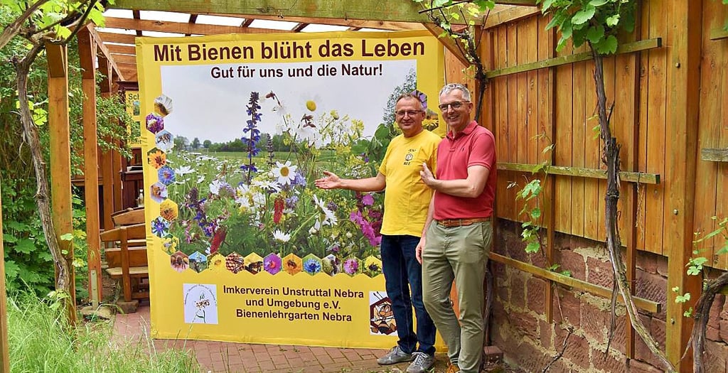 Vereinsvorsitzender Falko Breuer (l.) unterhält sich im Bienenlehrgarten vor einer Plakatwand zum Weltbienentag mit dem Naumburger Henning Jaworski.