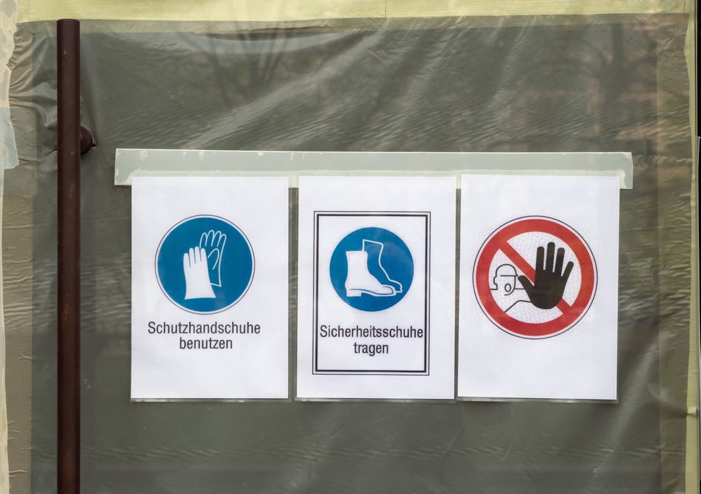 Schilder mit der Aufschrift für "Schutzhandschuhe benutzen", "Sicherheitsschuhe tragen" und dem Verbot für das Betreten der Baustelle hängen am Fenster Baustelle im Erdgeschoss eines Bürohauses.