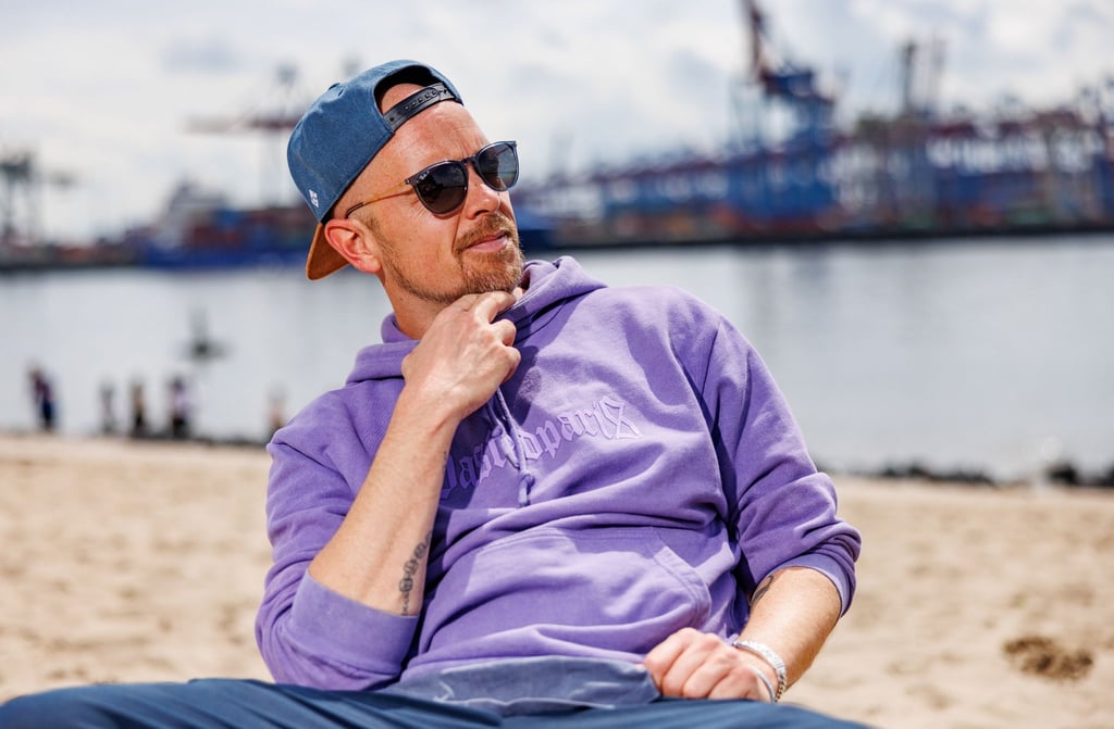 Der Musiker Jan Delay sitzt am Elbstrand in Hamburg vor den Industrieanlagen des Hafens in der Sonne.