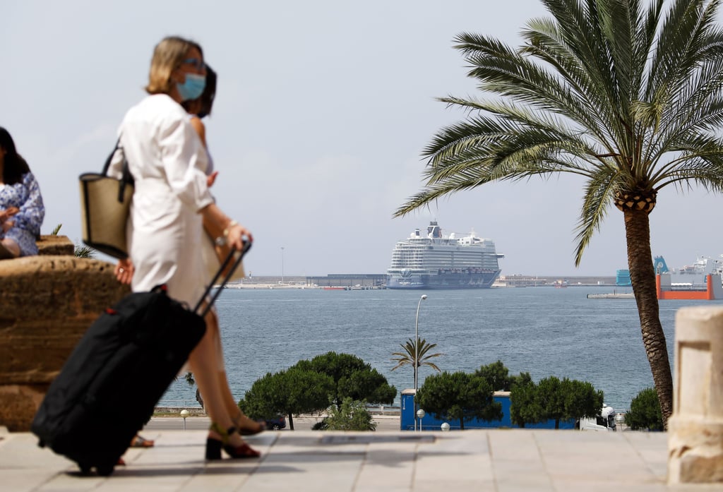 Symbolbild: Zwei Touristinnen gehen mit ihrem Gepäck über die Promenade von Palma de Mallorca gehen. Im Hintergrund liegt ein Kreuzfahrtschiff im Hafen der Baleareninsel.