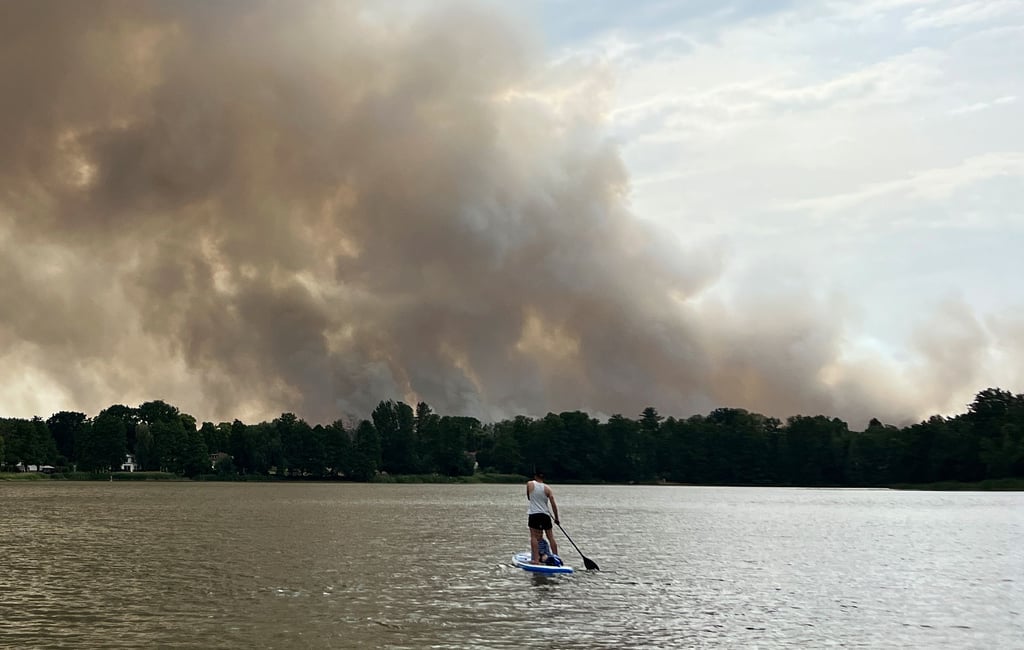 Ein junger Mann steht auf einem SUP auf dem Seddiner See, während eine Rauchwolke des Waldbrandes bei Treuenbrietzen weithin sichtbar ist.