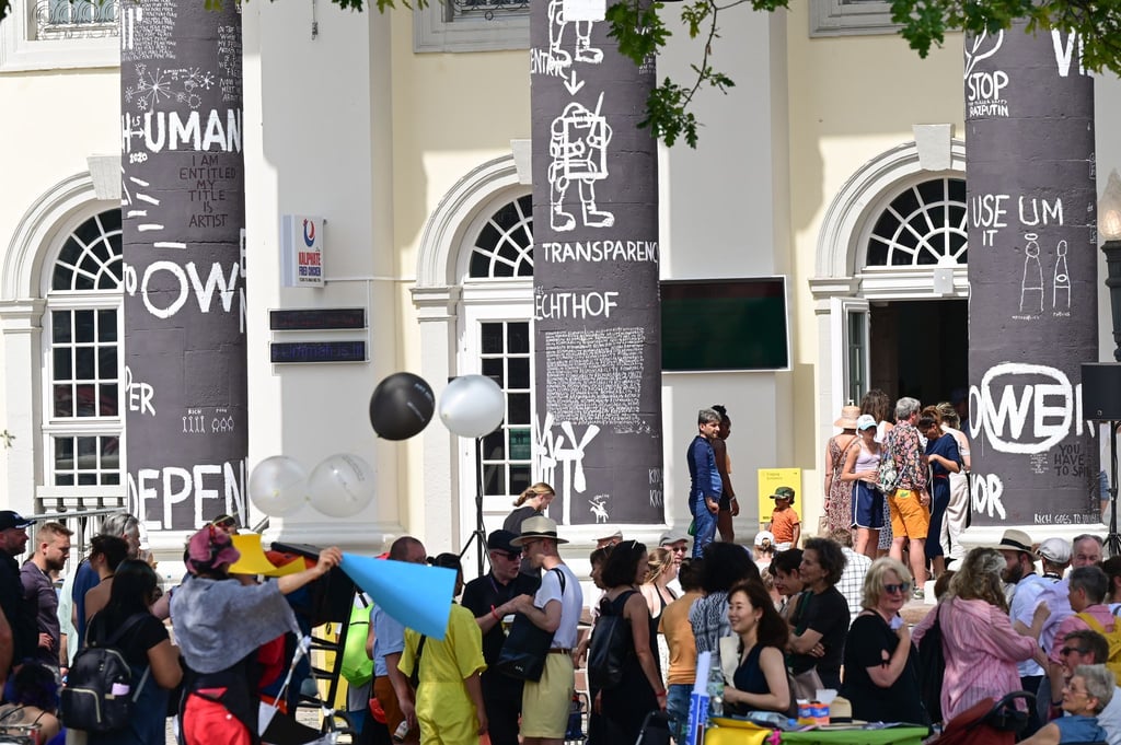 Besucher der documenta fifteen stehen nach Eröffnung vor dem Fridericianum, dessen Säulen der rumänische Künstler Dan Perjovschi mit schwarzer Farbe bemalt und mit weißen Symbolen und Zeichen zu Themen wie Frieden, Solidarität oder Nachhaltigkeit beschriftet hat.