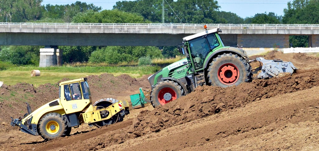 Ein Traktor mit angehängter Fräse vermischt vorn das alte Erdmaterial für den Deichkörper, dahinter walzt eine Schaffußwalze alles wieder fest. Im Hintergrund ist die Bahnbrücke zu sehen.