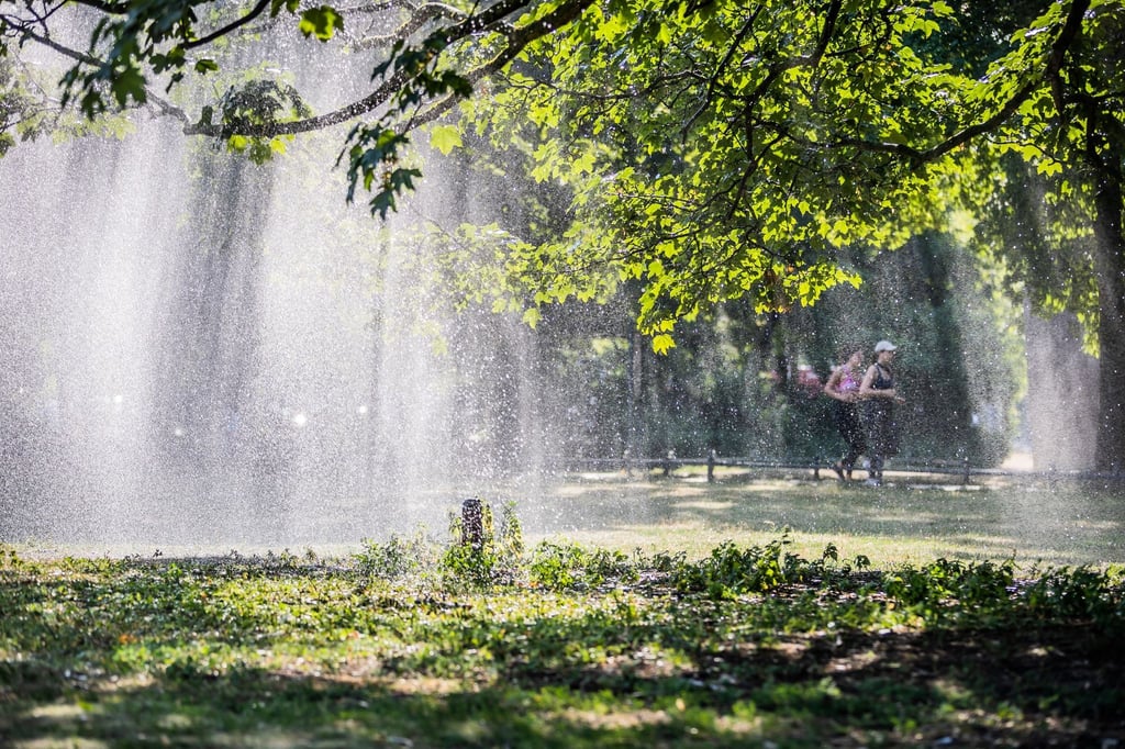 Zwei Joggerinnen laufen am Morgen hinter dem Wasserstrahl eines Rasensprengers im Berliner Tiergarten.