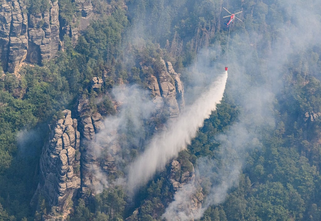 Ein Lastenhubschrauber wirft Löschwasser über einem Wald im Nationalpark Sächsische Schweiz ab.