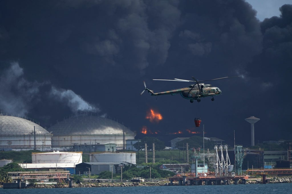 Ein mit Wasser beladener Hubschrauber fliegt über die Supertanker-Basis Matanzas, von der dunkler Rauch und Flammen aufsteigen, während Feuerwehrleute versuchen, den Brand zu löschen.