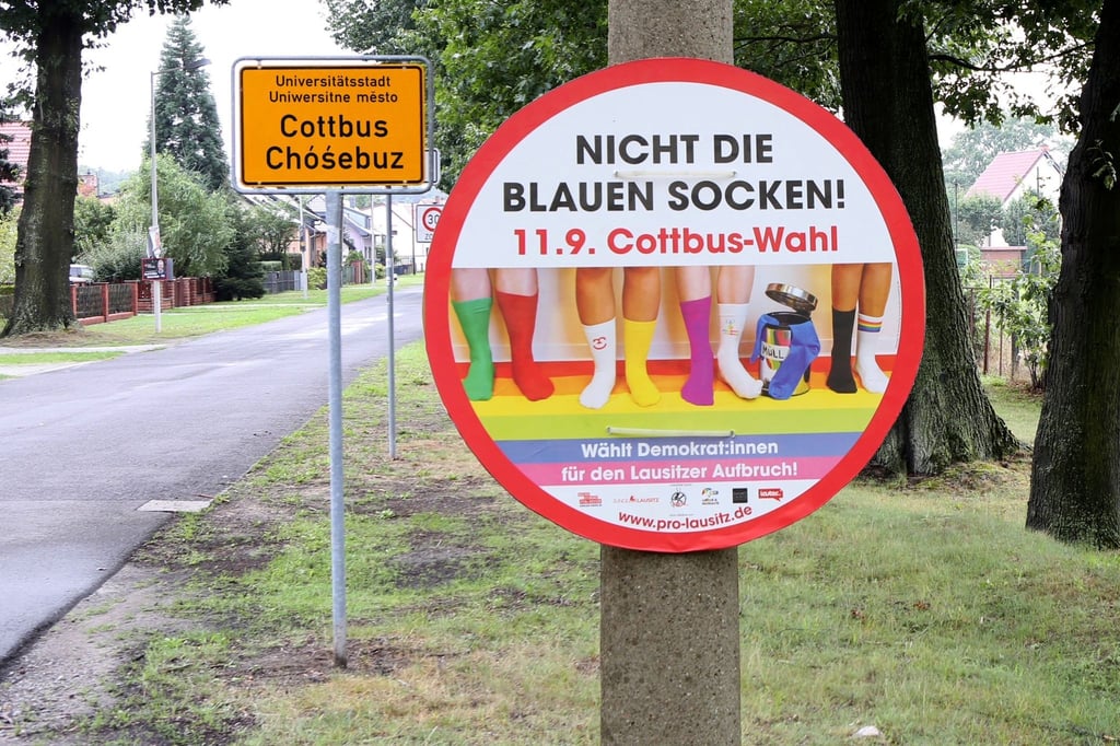 Vor dem Ortseingangsschild der Stadt Cottbus hängt an einem Pfahl ein Plakat der Kampagne „Blaue Socken“.