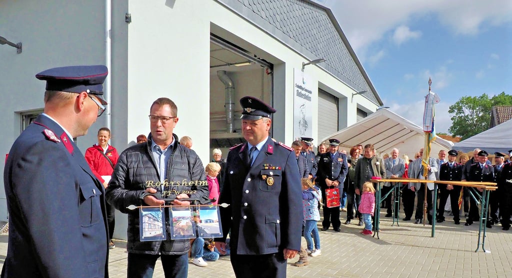 Fotos des Grundstücks vom Früher und Jetzt hängen am symbolischen Schlüssel, den Ortsbürgermeister Hans-Jörg Gifhorn (Mitte) und Bürgermeister Dirk Heinemann (rechts) an Ortswehrleiter Felix Böhm (links) vor dem neuen Rohrsheimer Feuerwehrhaus überreichten.