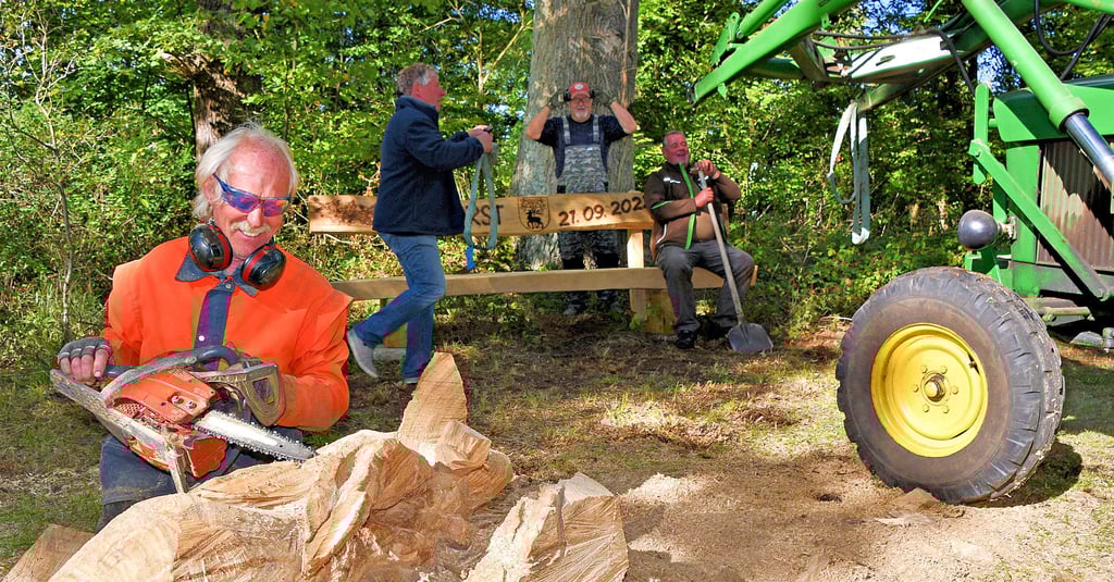 Lorenz Tacke aus Zobbenitz schnitzt mit der Motorkettensäge im Park in Dorst zwei Hausen aus dem Baumstamm. Im Hintergrund stellen Dorster die neue Bank am Teich auf.