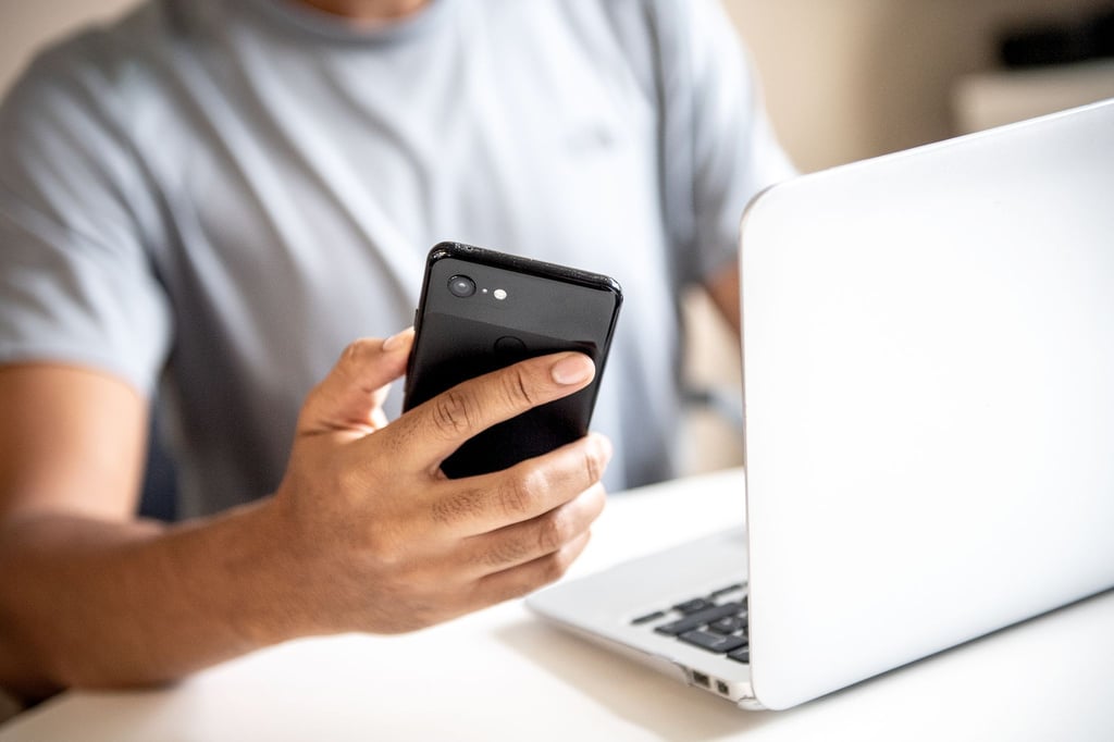 Ein Mann sitzt vor einem Laptop und hält ein Handy in der Hand.