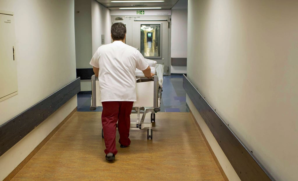 Fachpersonal wie dieser Pfleger, der einen Patienten durch einen Flur schiebt, fehlt - obwohl laut Statistik in den zwölf Jahren bis 2020 immer mehr Menschen im Gesundheitswesen gearbeitet haben.