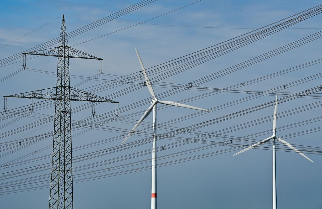 Windenergieanlagen stehen hinter Hochspannungsmasten von mehreren Stomleitungen.