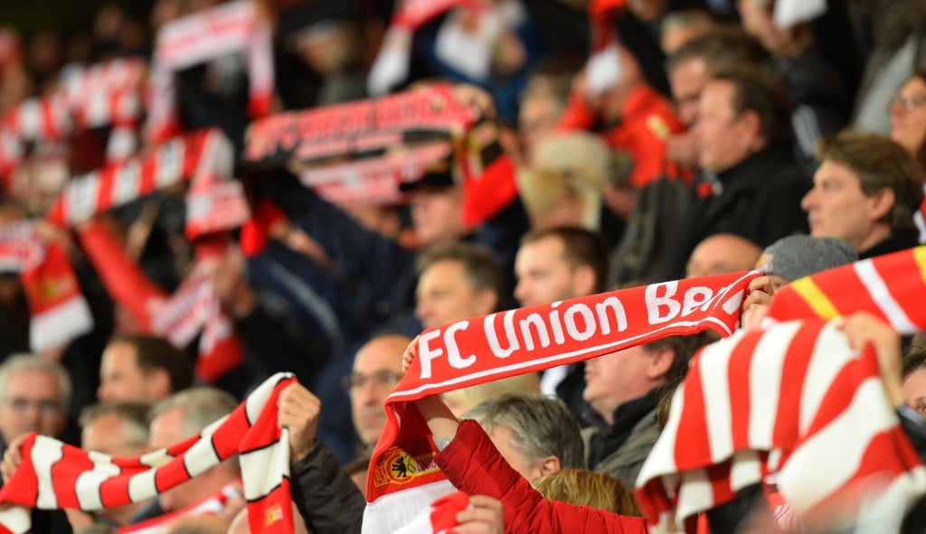 Fans des 1. FC Union Berlin halten Schals mit dem Vereinslogo in die Höhe.