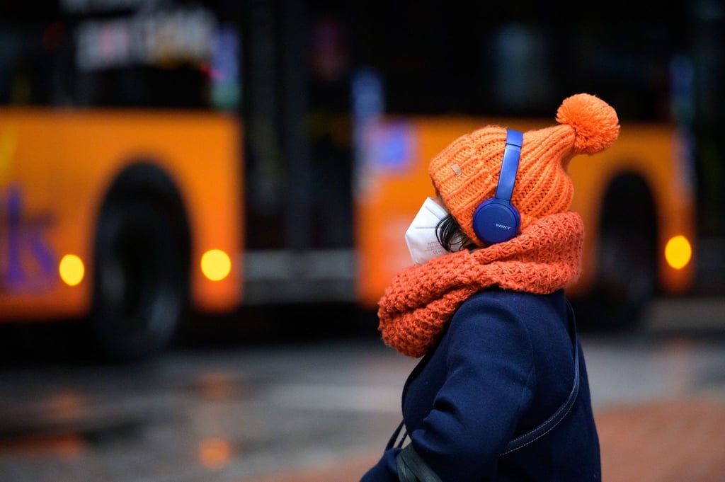 Eine Frau sitzt mit FFP2-Maske, dicker Mütze und Kopfhörern an einem Busbahnhof.