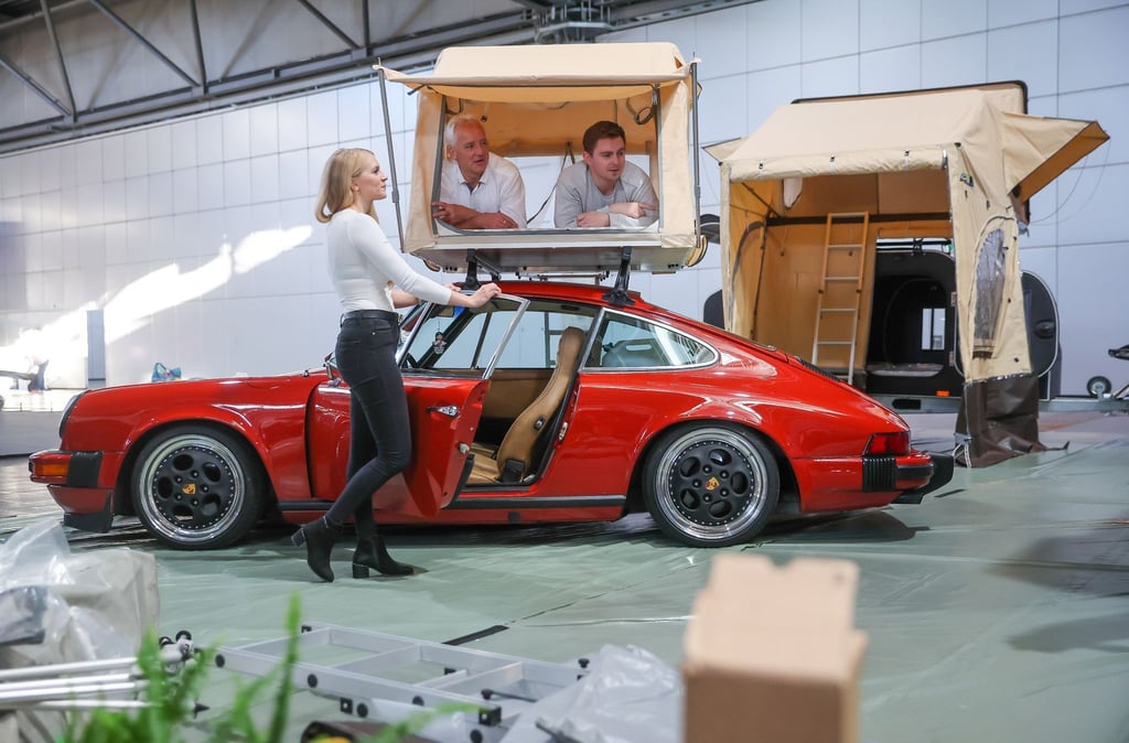 Zwei Männer und eine Frau posieren in einem Dachzelt auf einem Porsche in der Messehalle.