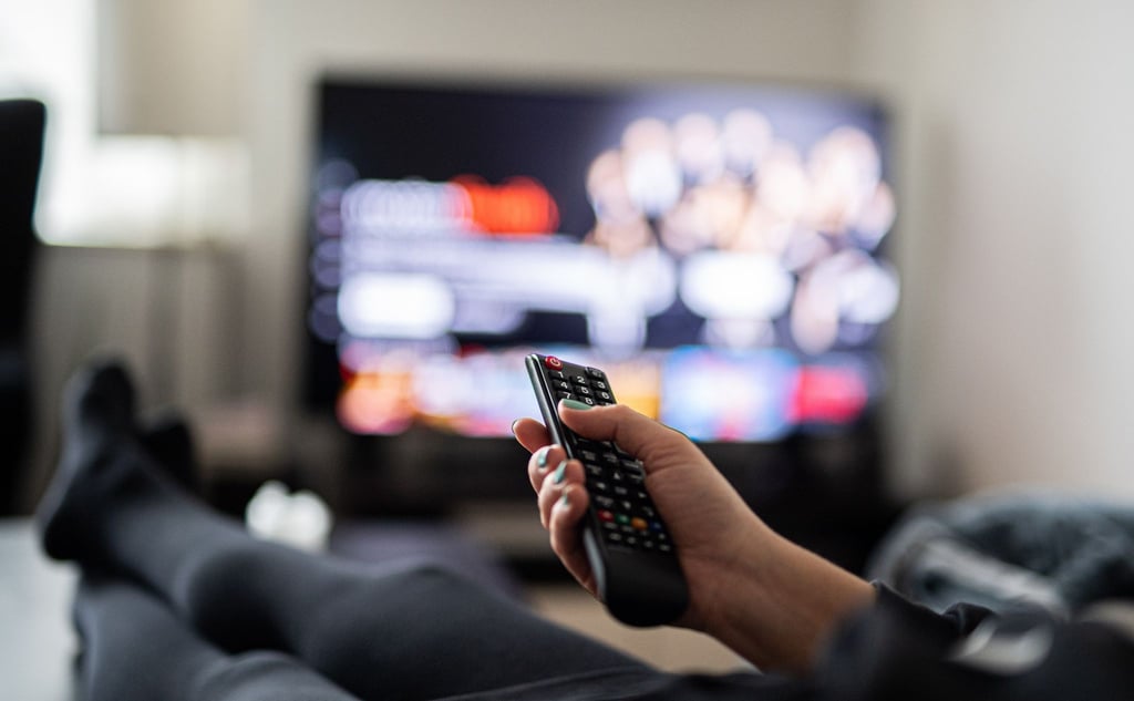 Eine Frau sitzt vor dem Fernseher und hat eine Fernbedienung in der Hand.