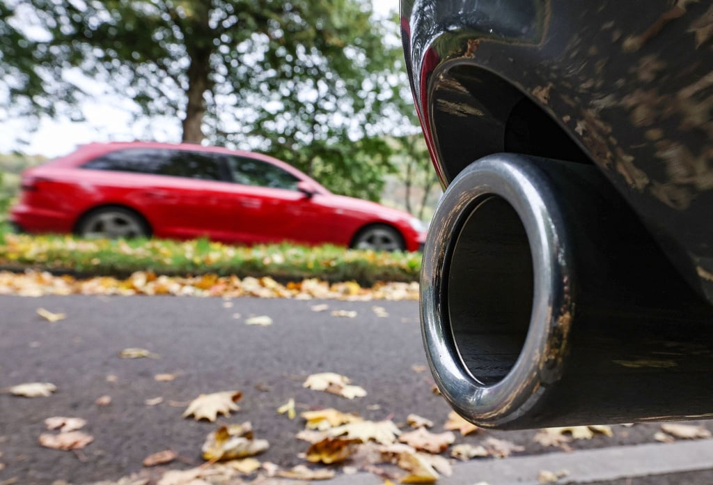 Hinter dem Auspuff eines Pkw fährt ein Auto vorbei.