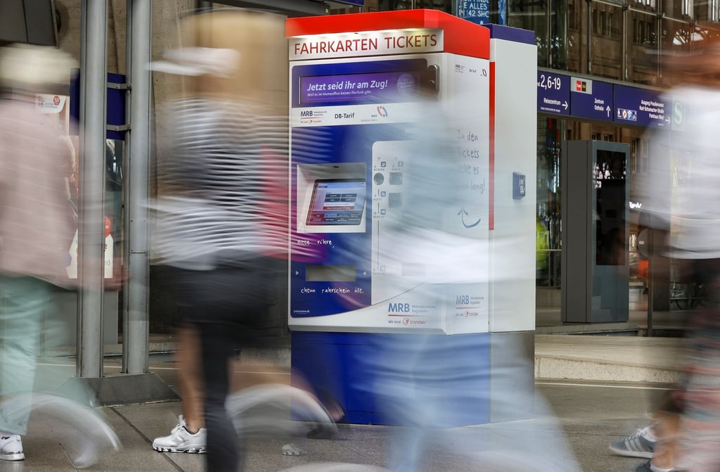 Passagiere gehen an einem Bahnhof an Fahrkartenautomaten vorbei.