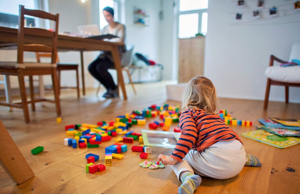 Ein zweijähriges Kind spielt im Wohnzimmer, während seine Mutter Zuhause im Homeoffice an einem Laptop arbeitet. Wer Homeoffice ermöglichen und sein Kind zu Hause betreuen kann, erweist derzeit vielen Kindergärten einen großen Dienst.