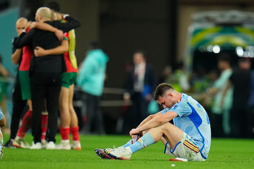 Spaniens Aymeric Laporte sitzt nach der Niederlage auf dem Spielfeld, während Marokkos Spieler im Hintergrund über ihren Sieg im Elfmeterschiessen jubeln.