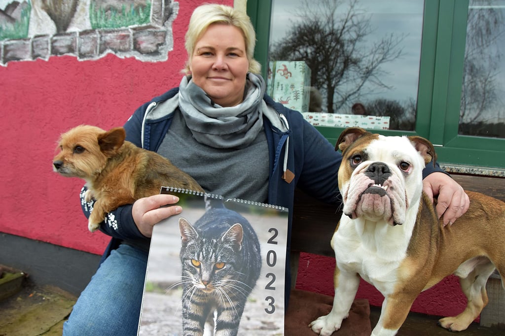 Tierheimleiterin Kathrin Behrends zeigt den Fotokalender 2023. Im Kalender, der für eine Spende zu haben ist, sind Bilder mit Tieren der Einrichtung abgedruckt. Seite an Seite mit der Chefin sitzen der Englisch-Bulldog-Rüde Georg und die Mischlingshündin Mira.