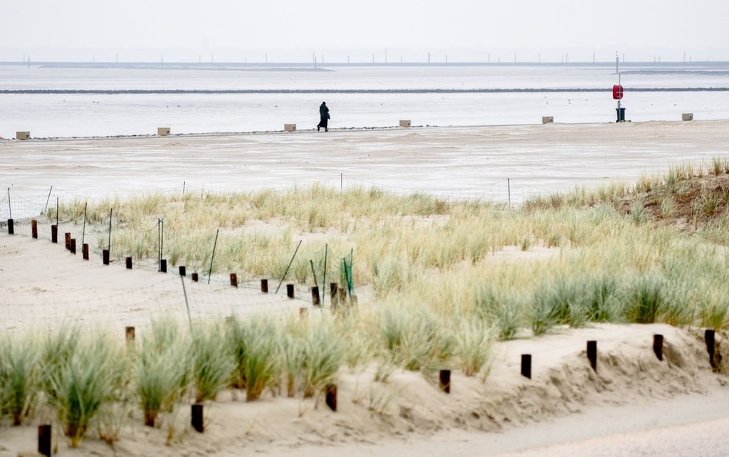 Eine Urlauberin geht bei regnerischem Wetter auf einem Weg am Strand vor der Nordsee entlang.