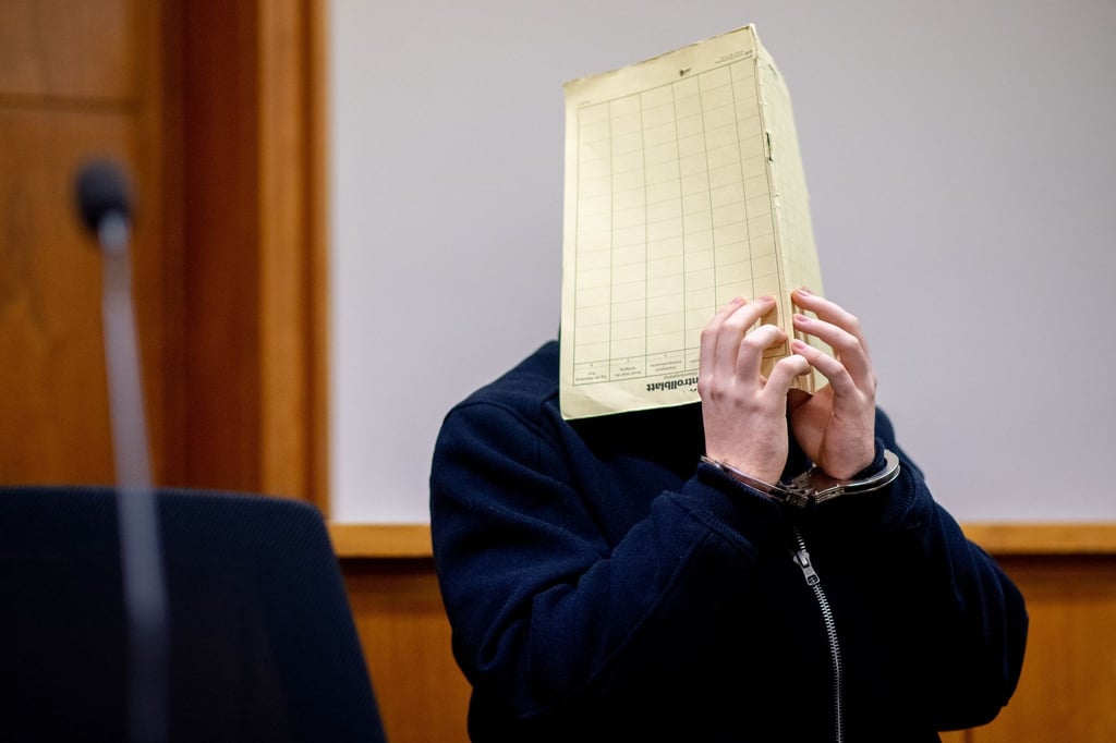 Der Angeklagte sitzt vor Prozessbeginn in einem Gerichtssaal im Landgericht und hält sich einen Aktendeckel vor das Gesicht.