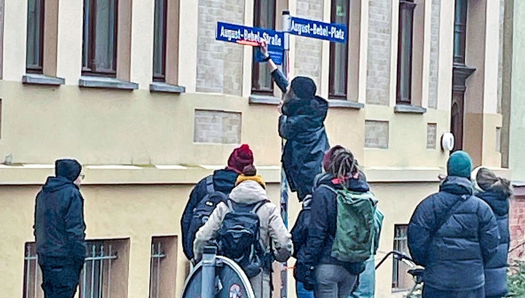 Eine Gruppe junger Menschen hat am August-Bebel-Platz in Halle das Straßenschild mit orangefarbenem Klebeband beklebt.