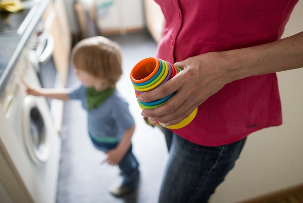 Eine Mutter hält Kinderspielzeug in der Hand, während ein kleiner Junge an einer Waschmaschine spielt. Die Krisen der letzten Jahre belasten vor allem Mütter - Stress und finanzielle Probleme sorgen für eine Dauerbelastung.